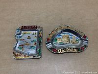 Two metal souvenir ashtrays placed side by side, the Arizona state shaped ashtray on the left and the Grand Canyon oval ashtray on the right.