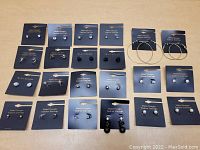 22 pairs of new fashion earrings arranged on black display cards, showing a variety of stud and hoop designs including pearls, rhinestones, and black stones.