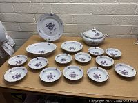 Lot of 17 porcelain dishes including a covered soup tureen, large serving platter, serving plate, and multiple smaller plates. All have a white base with purple floral decoration and scalloped edges.