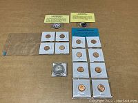 Photo showing an overview of the lot with coins in cardboard holders arranged in rows, two round medallions with yellow informational cards, and one larger silver-colored medallion in a protective plastic case.