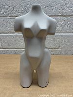 White ceramic female torso bust sculpture with smooth matte finish and stylized curves, displayed against a brick wall background.