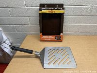 Photo showing the spatula outside of its original cardboard packaging with 'Mr. BBQ Stainless Steel Oversized Spatula' text visible on packaging behind it.