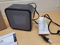 Small black cube-shaped desktop space heater with silver mesh grille, black power cord with fuse attached, placed on a table against a brick wall.