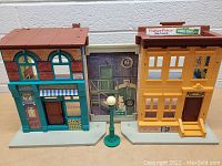 Front view of both Sesame Street buildings connected with street lamp between them on a brown surface against a white brick wall.