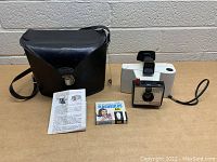 Photo of the Polaroid Swinger Land Camera Model 20 with wrist strap, black carrying case with shoulder strap, instruction sheet, and box of AG1 flashbulbs on a concrete background.