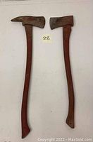 Two vintage fireman axes side by side with red wooden handles and metal heads on a plain surface.