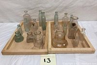 Full lot of 16 antique and vintage glass collectible bottles arranged in two wooden trays on a white surface.