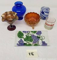 Photo showing all seven colored glass items in the lot, including blue vase, carnival glass comport, amber bowl, fused grape and vine glass tray, Harvard measuring cup, blue and white swirl glass, and purple stemmed glass