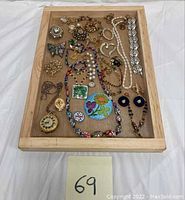 Overhead view of wooden tray holding assorted costume jewelry including necklaces, earrings, brooches, and bracelets for sale.