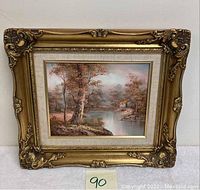 Full view of the framed oil painting showing the landscape scene with cabin, water, and trees.