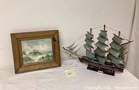 Photo of the framed beachscape painting and the wooden clipper model ship side by side on a white tablecloth, showing scale and condition of each.