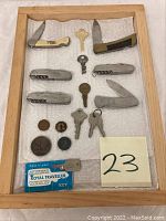 Full collection showing all items including 7 keys, 4 pocket knives, penny and tokens laid out on cloth inside wooden frame