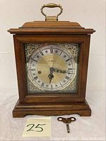 Front view of the vintage wooden mantel clock with decorative metal dial, hour and minute hands, and winding key shown in front.