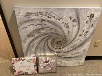Three art canvases leaning against wall. Large spiral textured canvas in neutral tones. Smaller floral and text canvases in front.