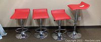 Four red adjustable bar stools with chrome bases placed side by side against a beige wall. Seats square with a slight curve, visible marks on the tops.