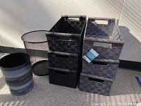 Photo showing six woven storage baskets stacked in two groups of three, three baskets in black and three in grey, all with wooden handles. Also visible are two trash bins; one plastic marbled grey and one black wire mesh.