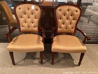 Front view of two matching chairs with tan upholstery and dark wood frames. Shows seat cushion, tufted backs, and carved arms.