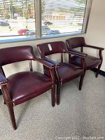 Three wood framed, maroon faux leather chairs with brass brad accents in armrest and seat edges, positioned in front of a window.