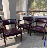 Three wood frame chairs with dark brown faux leather upholstery and brass stud detailing lined up in corner by windows