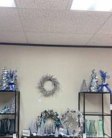 Overall display of silver and blue Christmas trees, wreaths, and vases on black shelving units against a beige wall.
