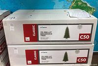 Two boxed 7-foot Christmas trees labeled '7ft Pre-Lit Braddock Tree' with '300 Clear Lights' and 42 inch diameter, stacked and shown in original packaging.