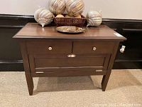 Front view of wood console table showing two drawers and larger drop-down storage compartment with round knobs and cup pull handle. Tapered legs and wood grain surface.