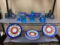 Shelf with three blue and white patterned bowls, nine blue glass bottles of various sizes, two sky blue Lex Water Goblets, and eight Gibson plates with two metal display stands beneath.