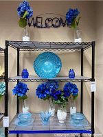 Shelving unit displaying six clear glass vases with white stones and blue florals, blue glass jars, turquoise bowl, candle holder, and swirl plates and bowls with metal Welcome sign above.