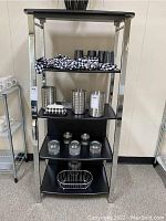 Four-tier metal and black shelving unit displaying various kitchen accessories including napkins, tumblers, utensil holders, jars, and a wire basket