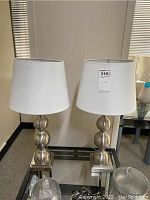 Pair of silver metal table lamps with white oval shades on glass table.