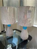 Two 22-inch tall new table lamps with ribbed ceramic bases and white fabric shades still wrapped in plastic on a glass top table, with window blinds background