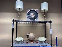 Two lamps with white shades on a black metal shelf along with a rose gold and black wall clock in box, a rose gold and pink globe, two white flower arrangements in boxes, and two rose gold unicorn figurines.