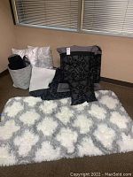 Photo of entire lot showing grey and black comforter set with pillows, basket with throw and pillows, and a white-grey shag rug.