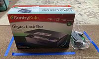 Box of SentrySafe large digital lock box with lock box partially visible next to it among tools and plastic basket.