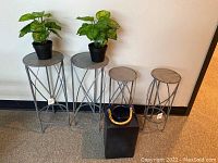 Four metal plant stands of two sizes with two faux plants in black pots and a black glass lantern with bamboo handle.