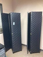 Photo of two three-panel black fiber weave room dividers standing side by side in a room corner, showcasing the woven texture and hinges.