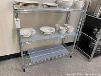 Front view of the metal wire shelving unit with white plates, bowls, and mugs on the top two shelves, bottom shelf empty.