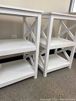 Two white laminate hardwood shelf units with three shelves and X-frame design shown side by side indoors on carpeted floor.