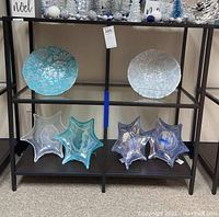 Shelving unit displaying glass and star-shaped decorative bowls in blue and clear hues on wood look bottom shelves and glass middle shelf with metal black frame.