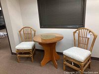 Full view showing a round particle board table with a glass top and two rattan chairs with white cushions placed around it in a room corner.