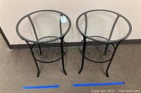 Two round metal and glass side tables with scrollwork base, placed side by side on carpeted floor.