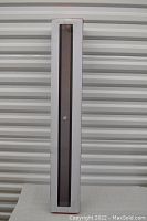 Photo showing the rectangular white box packaging containing the soundbar, with the black soundbar visible through a clear plastic window on the front.