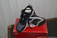 Black women's running shoes positioned on and beside a red New Balance branded box. Shoes have black and gray mesh with white cushioned soles.