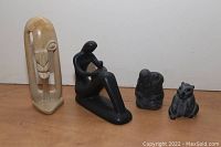 Four statues on wooden surface showing overall lot including tall beige abstract statue, black resin seated figure, dark stone person with fish, dark stone bear