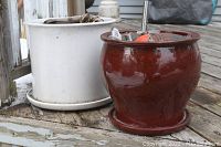 Two large ceramic planters side by side on wooden deck, a white one approximately 12 inches wide and a brown glossy one approximately 10 inches wide, both include saucers.