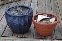 Photo showing blue glazed ceramic planter and terra cotta clay planter with broken pieces inside on wooden deck.