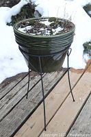 Full view of the green ceramic planter placed on a metal stand outdoors on a wooden surface with snow in the background.