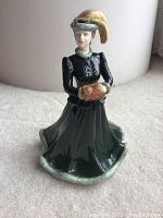 Porcelain figurine standing on carpeted floor showing full front and side view with dark green dress, black bodice, yellow plume hat, and fur muff.