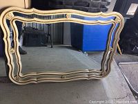 Large rectangular mirror with cream and gold detailed carved frame resting on floor in storage area.