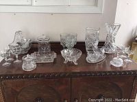 Wide view of miscellaneous crystal glassware collection on wooden cabinet, showing vases, glasses, bowls, and covered jars with varied patterns and sizes.